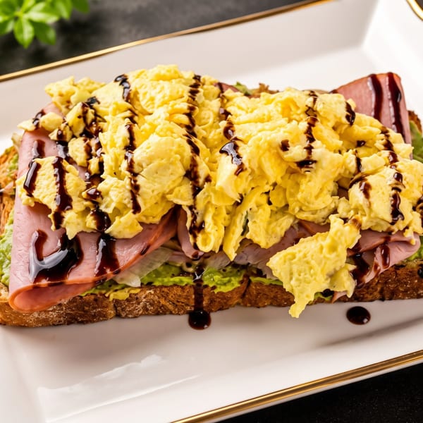 Scrambled eggs and ham on avocado toast, drizzled with balsamic glaze, on a white plate.